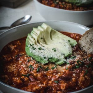turkey chili in a white bowl with sliced avocado and chives topping