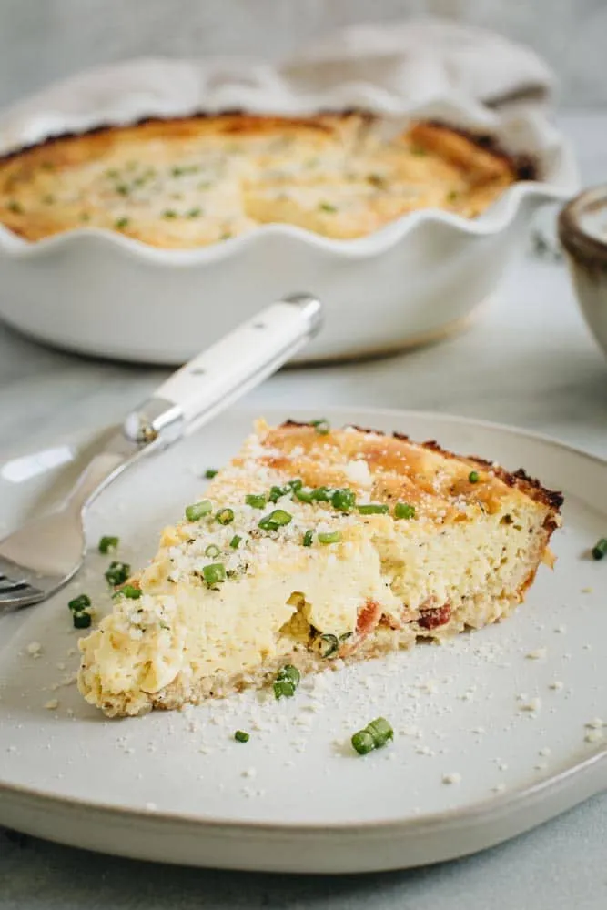 Cauliflower crust quiche slice topped with chives and grated cheese on white plate.