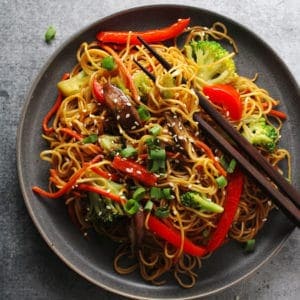 Vegetable lo mein on a black plate with brown chopsticks.