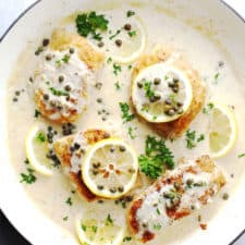 Creamy lemon cod piccata in a white skillet topped with lemon slices and fresh parsley.