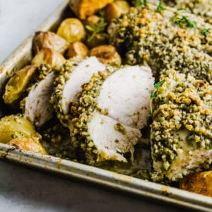 Walnut pesto chicken sliced on a rimmed baking sheet with potatoes surrounding it.