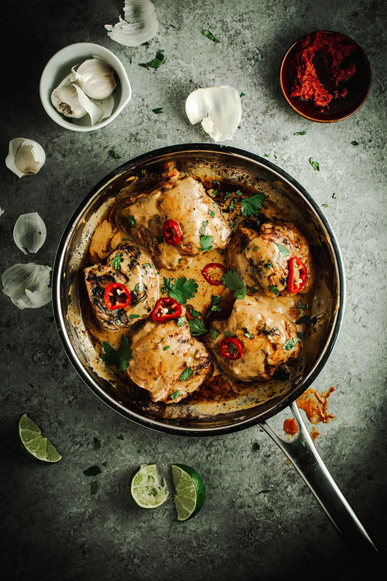 Easy Thai chicken topped with red chilies and fresh cilantro in a silver skillet with lime wedges and garlic surrounding it.
