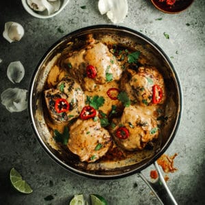 Easy Thai chicken topped with red chilies and fresh cilantro in a silver skillet with lime wedges and garlic surrounding it.