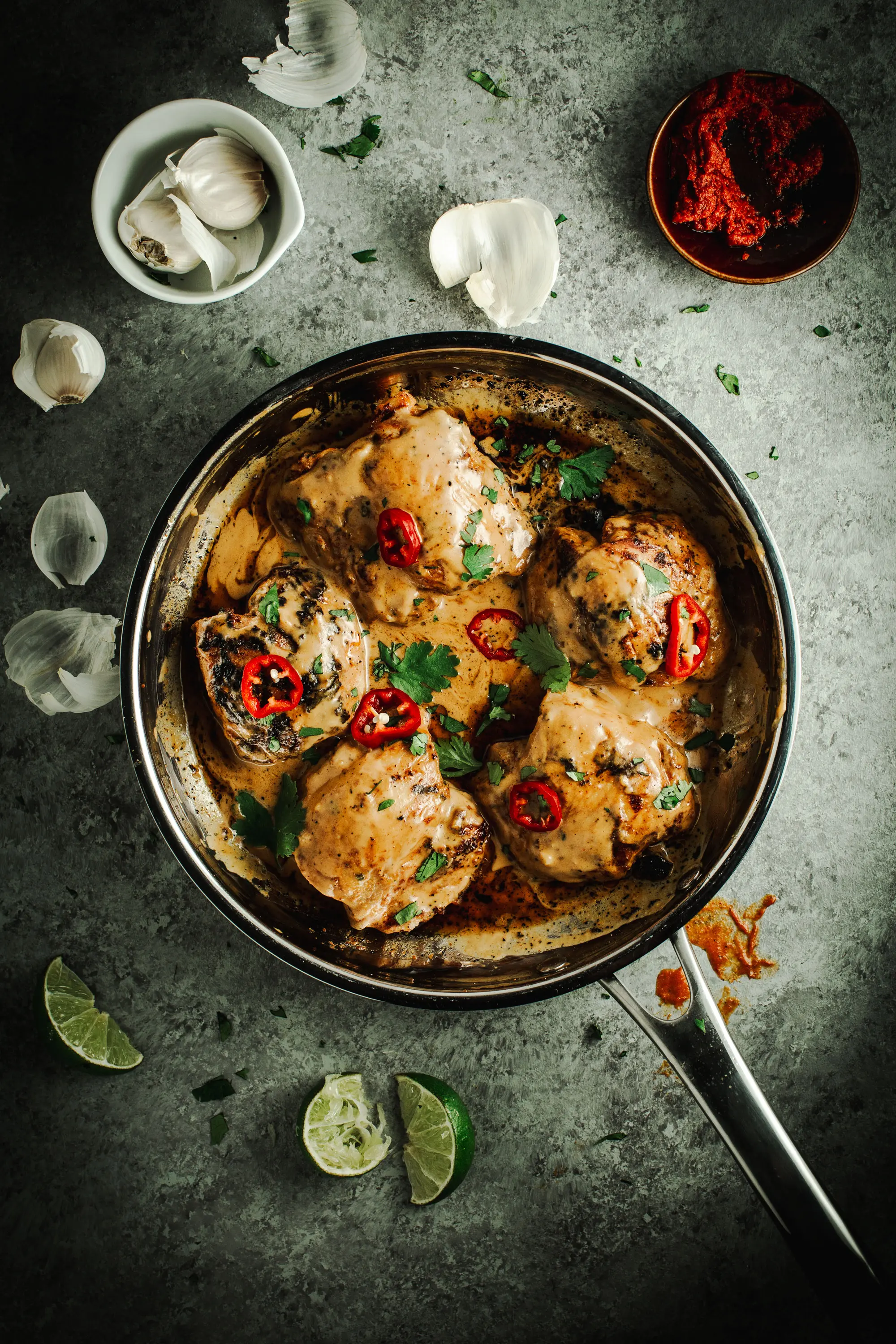 Easy Thai chicken topped with red chilies and fresh cilantro in a silver skillet with lime wedges and garlic surrounding it.