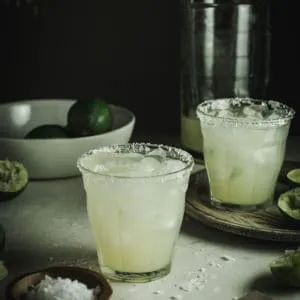 Margaritas in two glasses with salted rim and a bowl of limes behind.