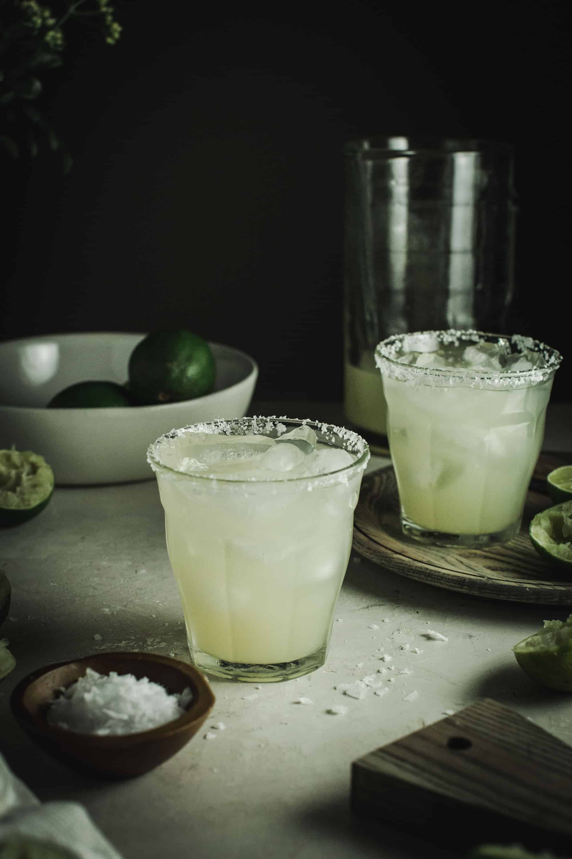 Margaritas in two glasses with salted rim and a bowl of limes behind.
