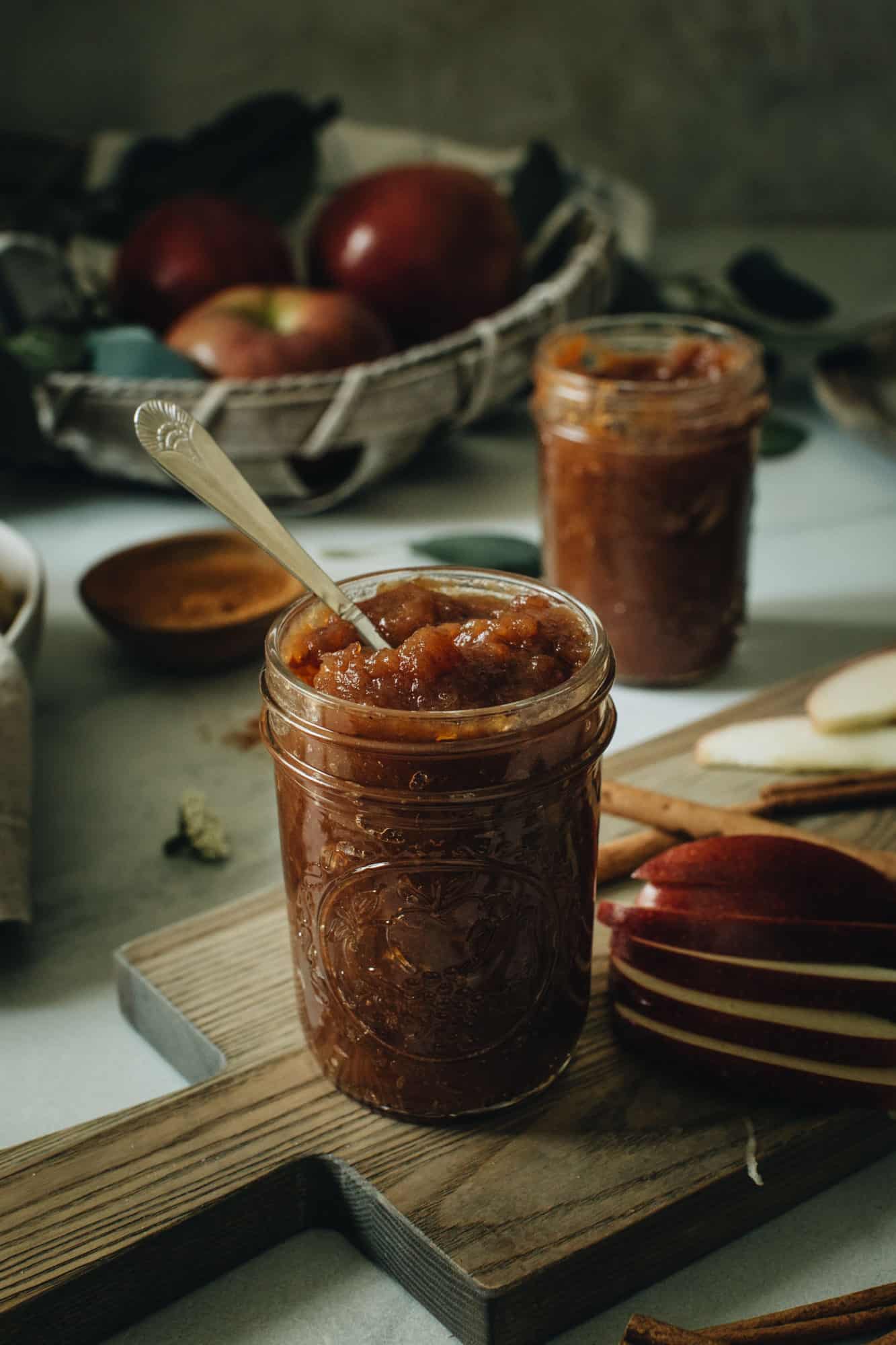 Cinnamon apple butter in a glass jar sitting on a wooden cutting board with a silver spoon stuck in the top.