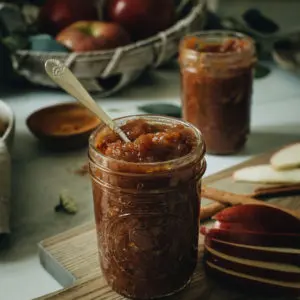 Cinnamon apple butter in a glass jar sitting on a wooden cutting board with a silver spoon stuck in the top.