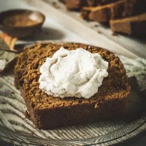 Moist pumpkin bread slice topped with whipped cream on a wooden plate.