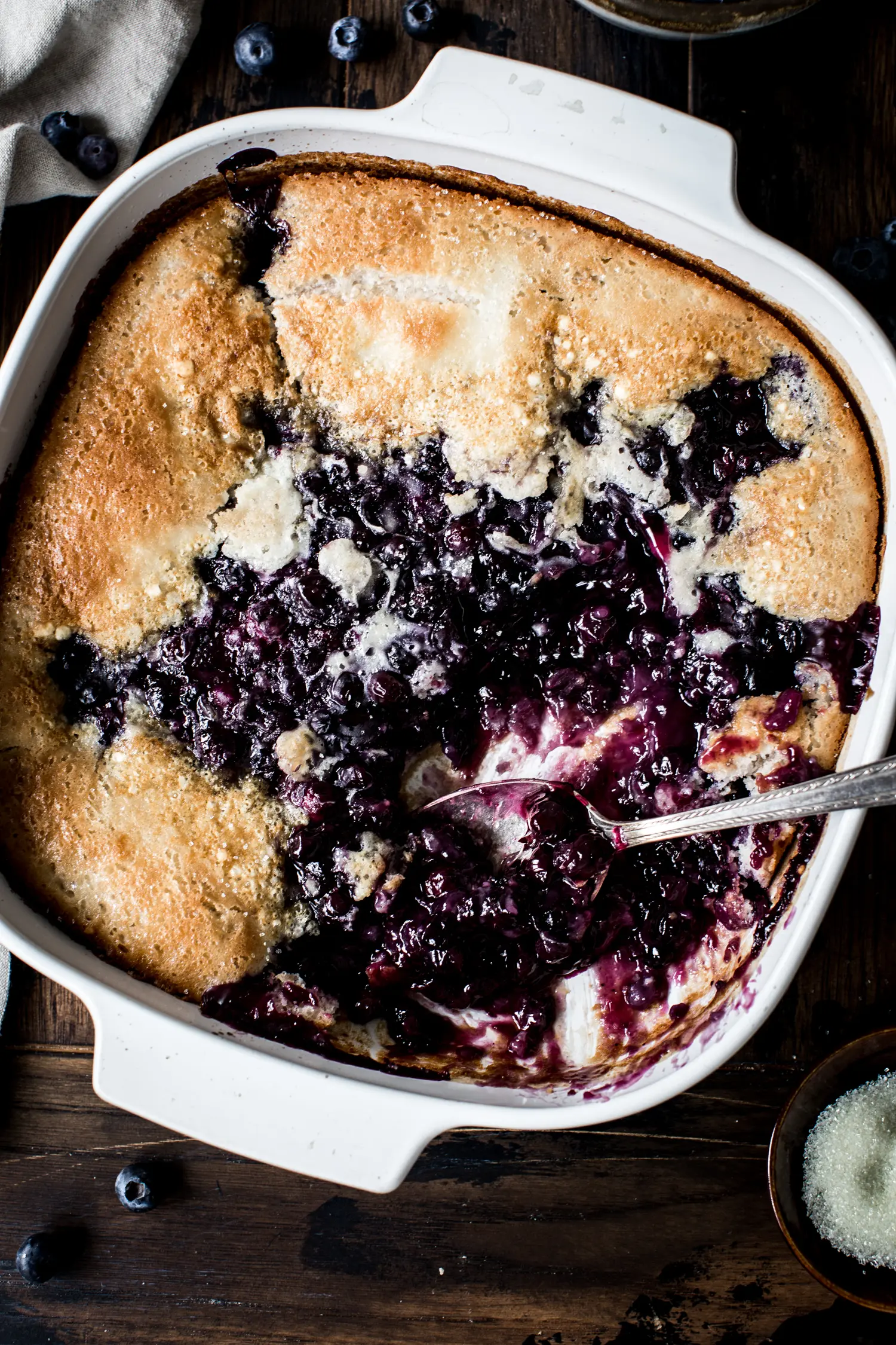 Close up of blueberry cobbler in white baking dish with silver spoon scooping it out.