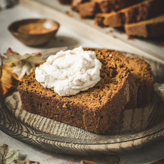 Slice of pumpkin bread topped with whipped cream on a wooden plate.