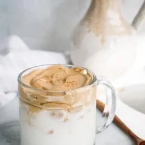 Whipped coffee in a clear glass mug with carafe in the background.