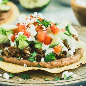 Ground beef tostada with toppings of lettuce, tomato, and crumbled cheese on a wooden plate.