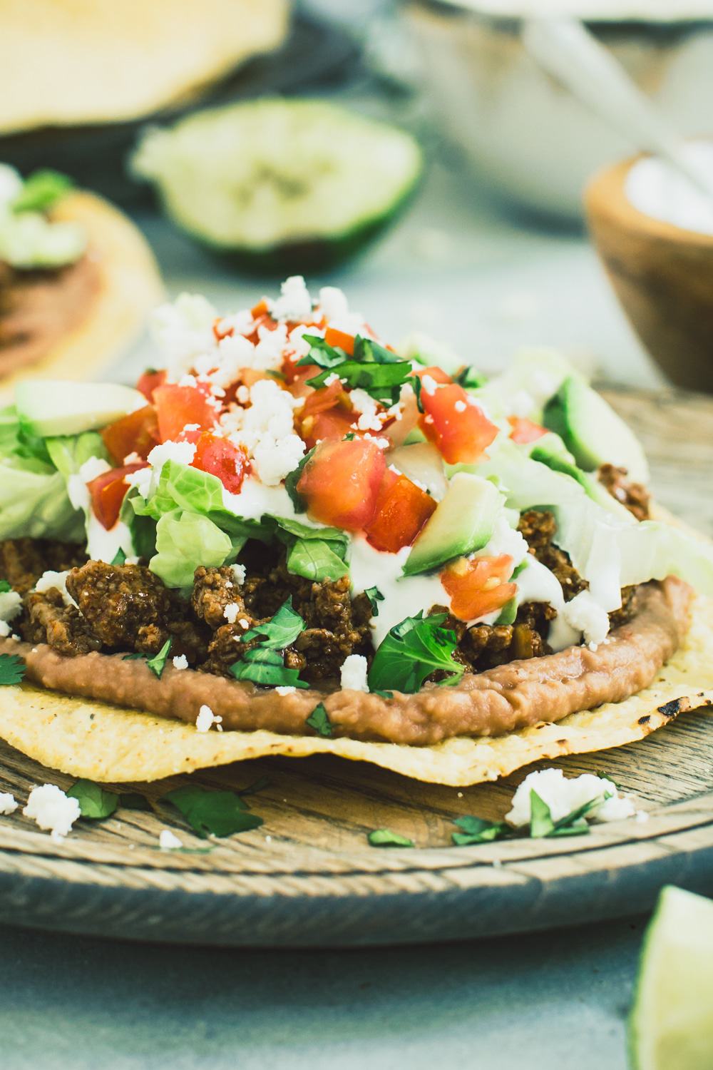 Ground beef tostada with toppings of lettuce, tomato, and crumbled cheese on a wooden plate.