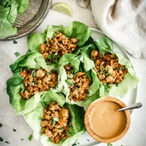 Asian chicken lettuce wraps on white round plate with peanut sauce in wooden bowl