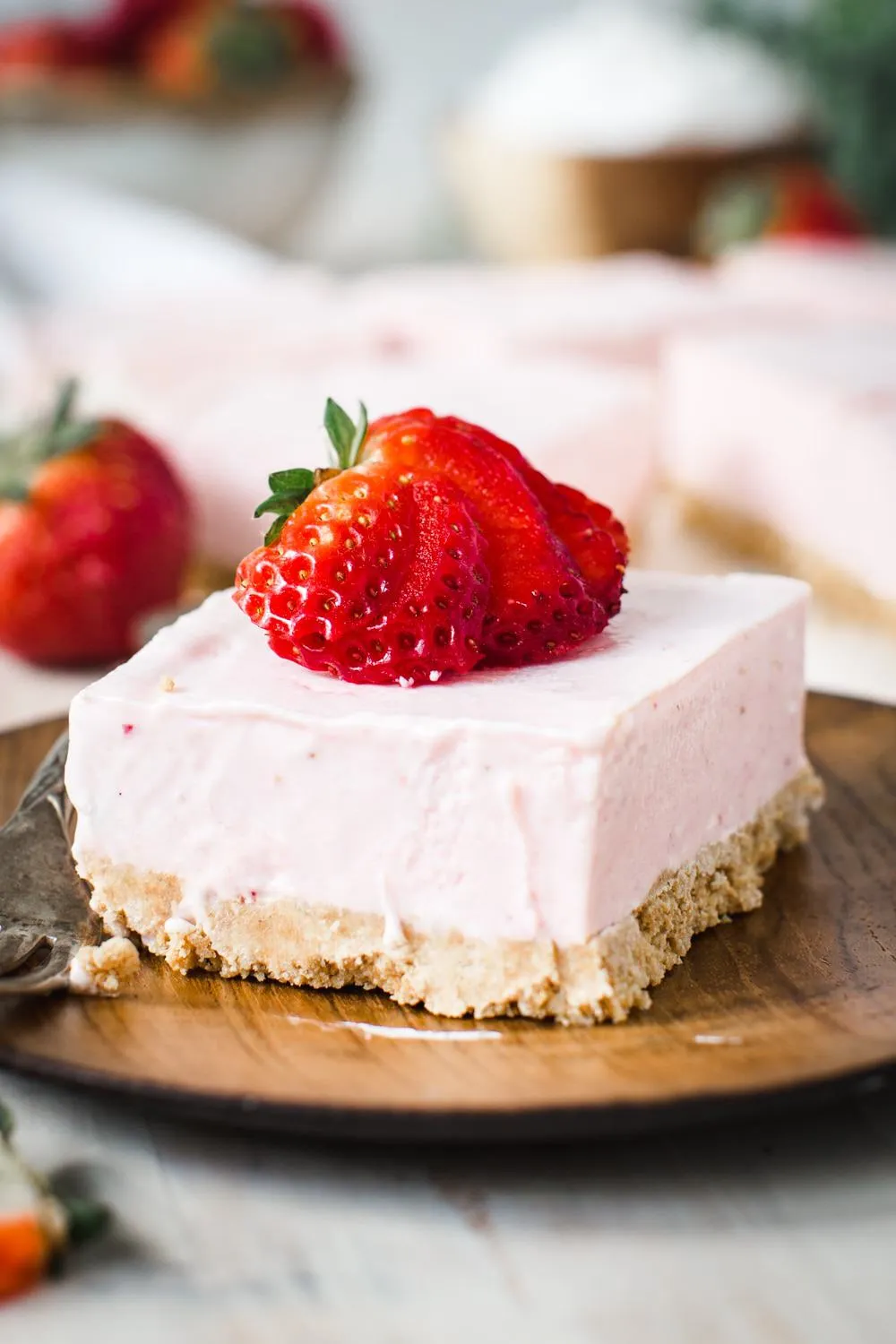 Square of strawberry cheesecake dessert topped with sliced strawberry on wooden plate.