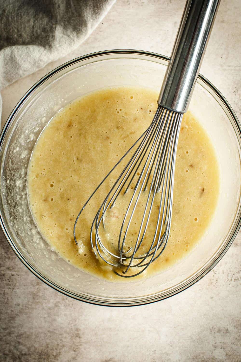 Banana bread ingredients mixing in a bowl with a wire whisk.