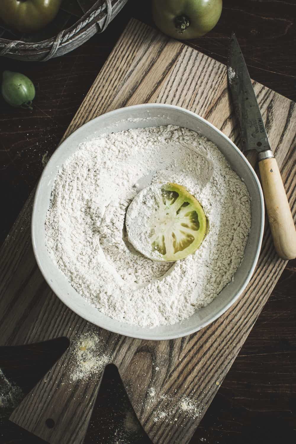 Flour mixture in a shallow white bowl with green tomato.