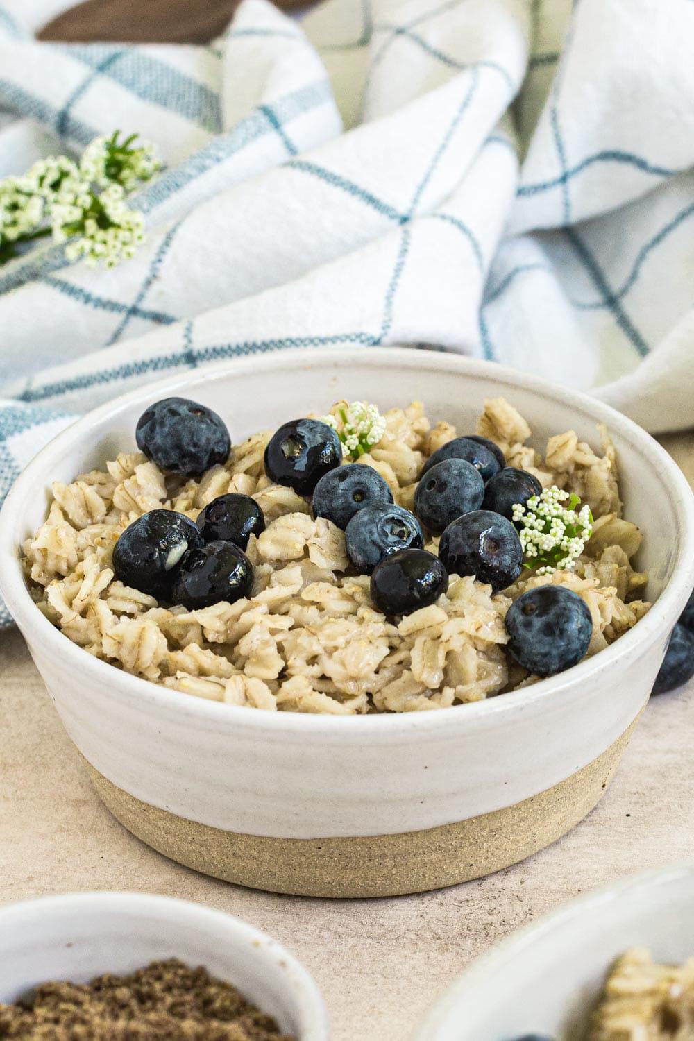flaxseed oatmeal topped with blueberries and syrup in a white bowl with blue checked dish towel behind
