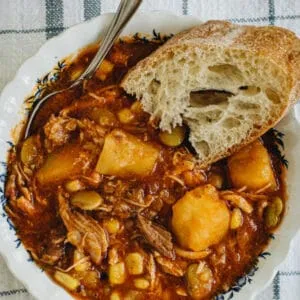 Brunswick Stew in a white and blue bowl with spoon and bread slice. White title with orange background.