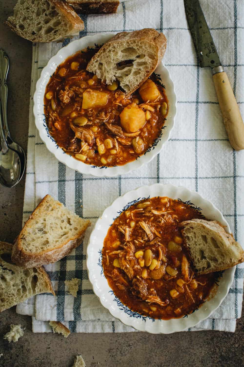 Two bowls of Alabama Camp Stew with bread slices on a white and blue checked towel.
