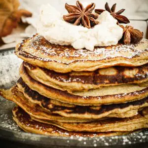 Pumpkin pancakes stacked and covered in powdered sugar and topped with whipped cream.