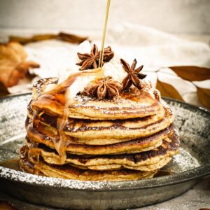 Syrup drizzling on top of pumpkin pancakes topped with whipped cream, powdered sugar, and star anise.