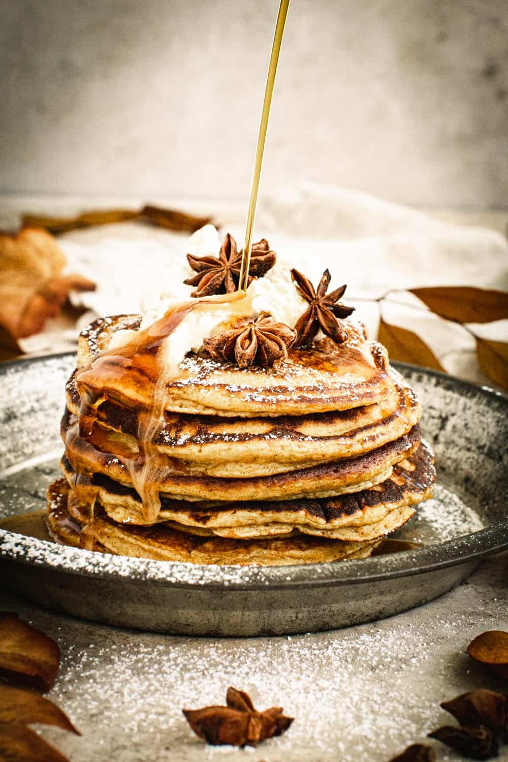 Syrup drizzling on top of pumpkin pancakes topped with whipped cream, powdered sugar, and star anise.