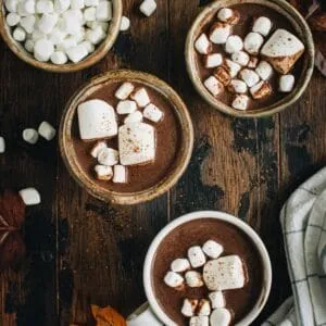 Three cups of pumpkin hot chocolate topped with marshmallows next to a bowl of mini marshmallows.