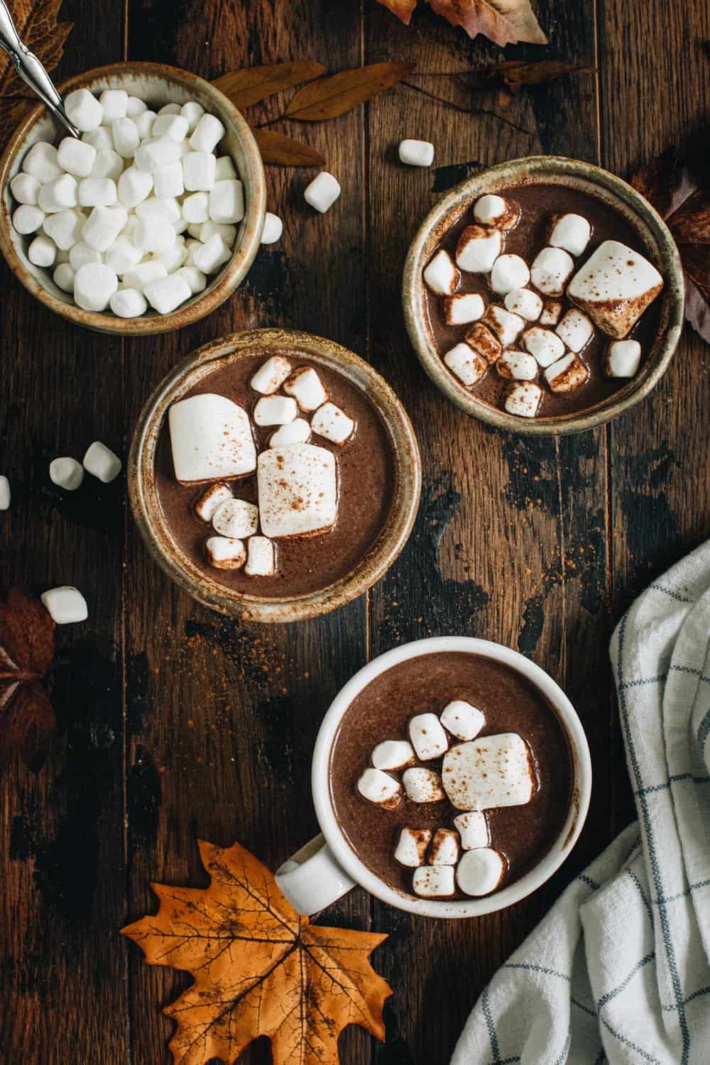 Three cups of pumpkin hot chocolate topped with marshmallows next to a bowl of mini marshmallows.