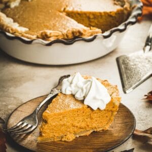 Slice of pumpkin pie on a wooden plate with a silver fork.