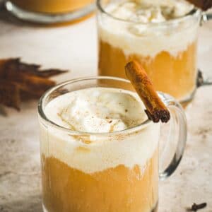 Hot bourbon apple cider cocktail topped with whipped cream and a cinnamon stick in a glass mug.