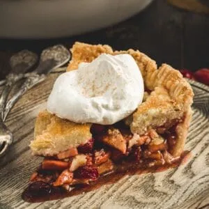 Slice of apple and cranberry pie topped with whipped cream and sitting on a wooden plate.