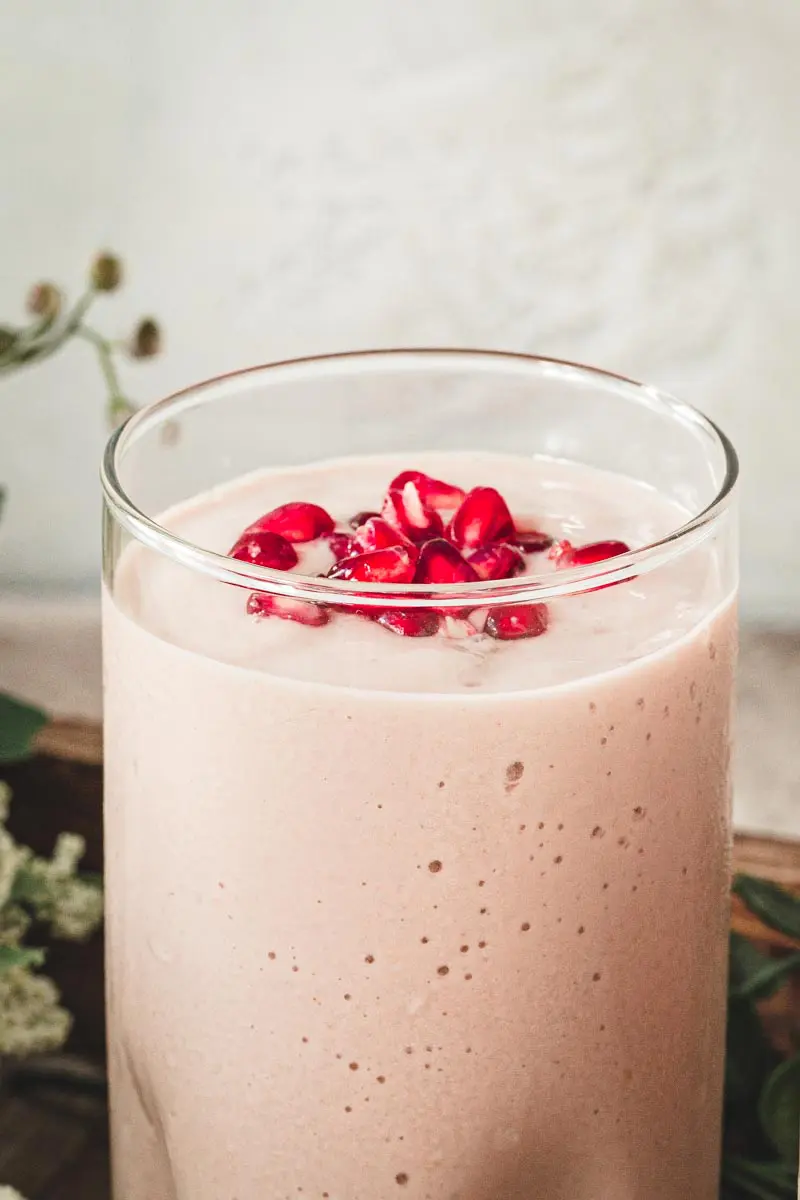 Pink colored smoothie in a tall glass with pomegranate seeds on top.