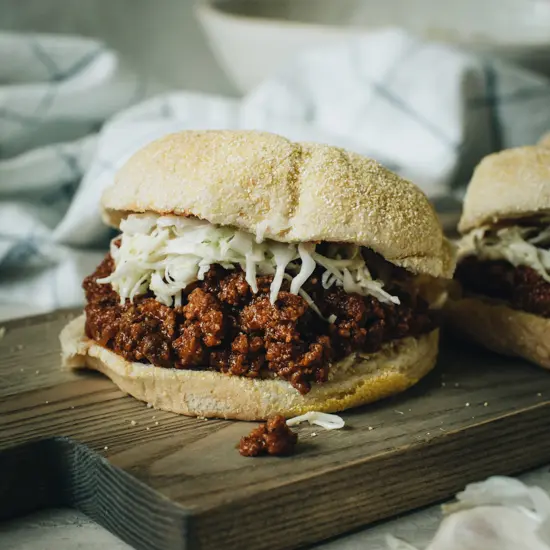 Sloppy Joe topped with coleslaw on a seeded bun sitting on a wooden cutting board.