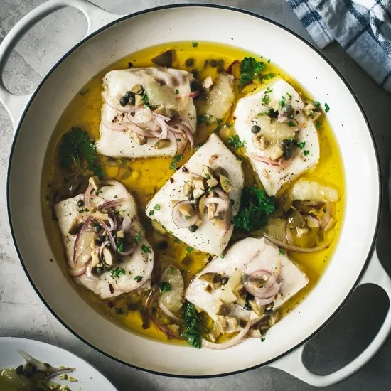 Cod in marinade topped with onion and olive relish in white skillet.