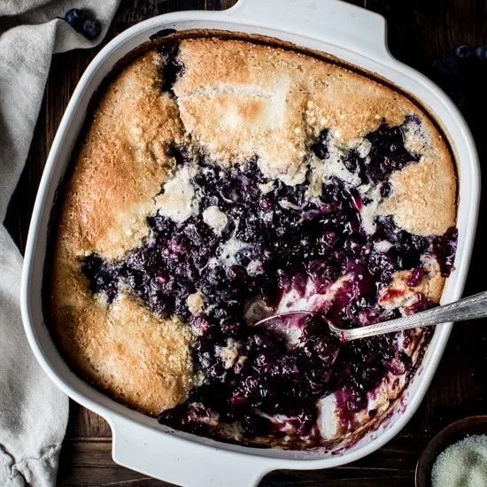 Blueberry cobbler in a square baking dish with a silver serving spoon.