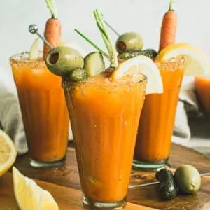 Carrot Juice Bloody Mary cocktails in glasses topped with garnishes and sitting on a wooden board.