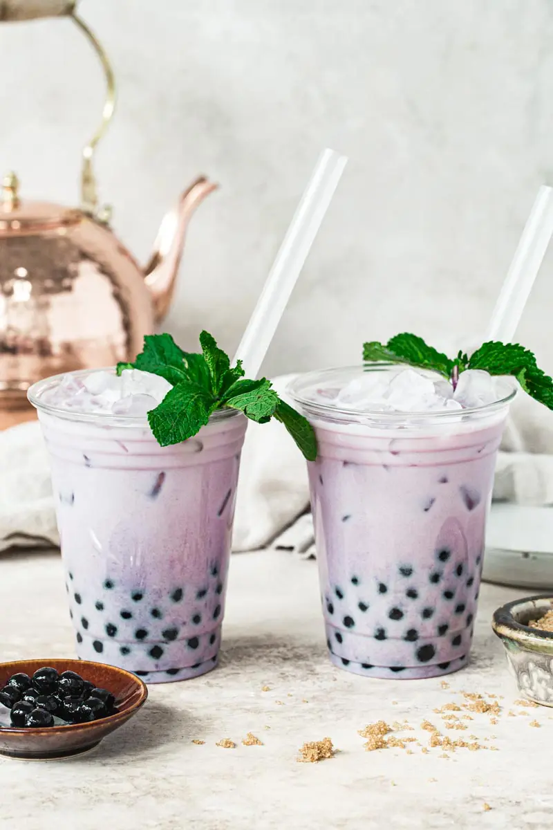 Two taro bubble teas in plastic cups topped with mint and tapioca pearls in a small dish next to them.