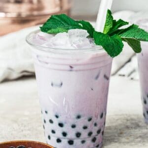Taro milk tea in a plastic cup topped with mint leaves and a wide straw and a bowl of tapioca pearls next to it.