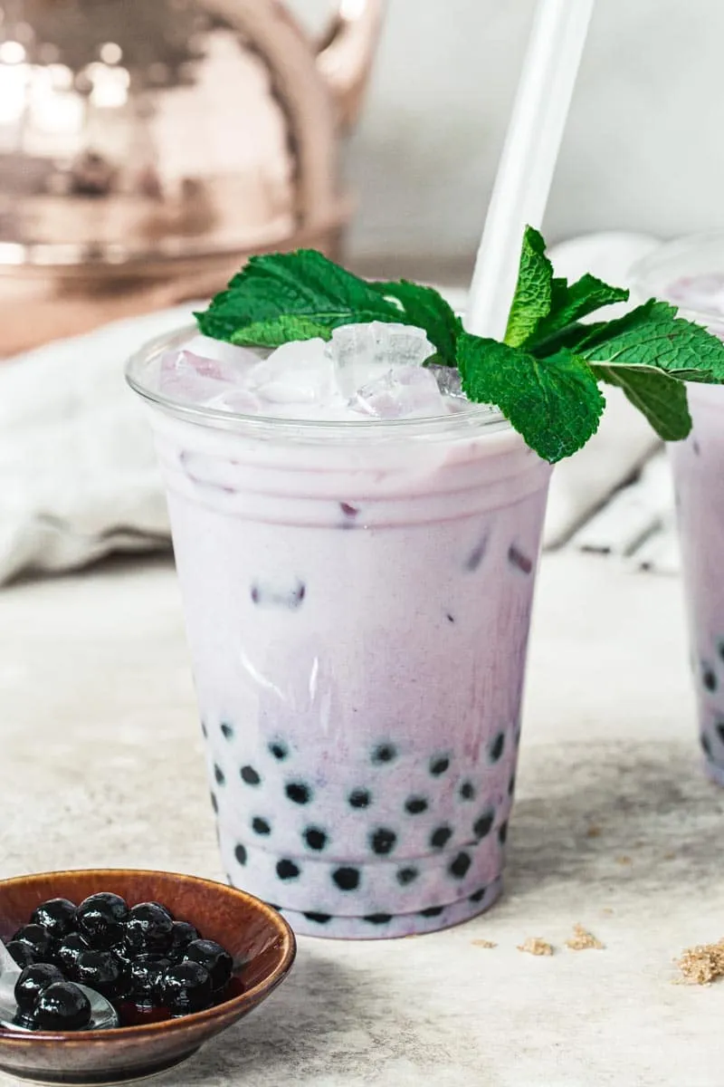 Taro milk tea in a plastic cup topped with mint leaves and a wide straw and a bowl of tapioca pearls next to it.