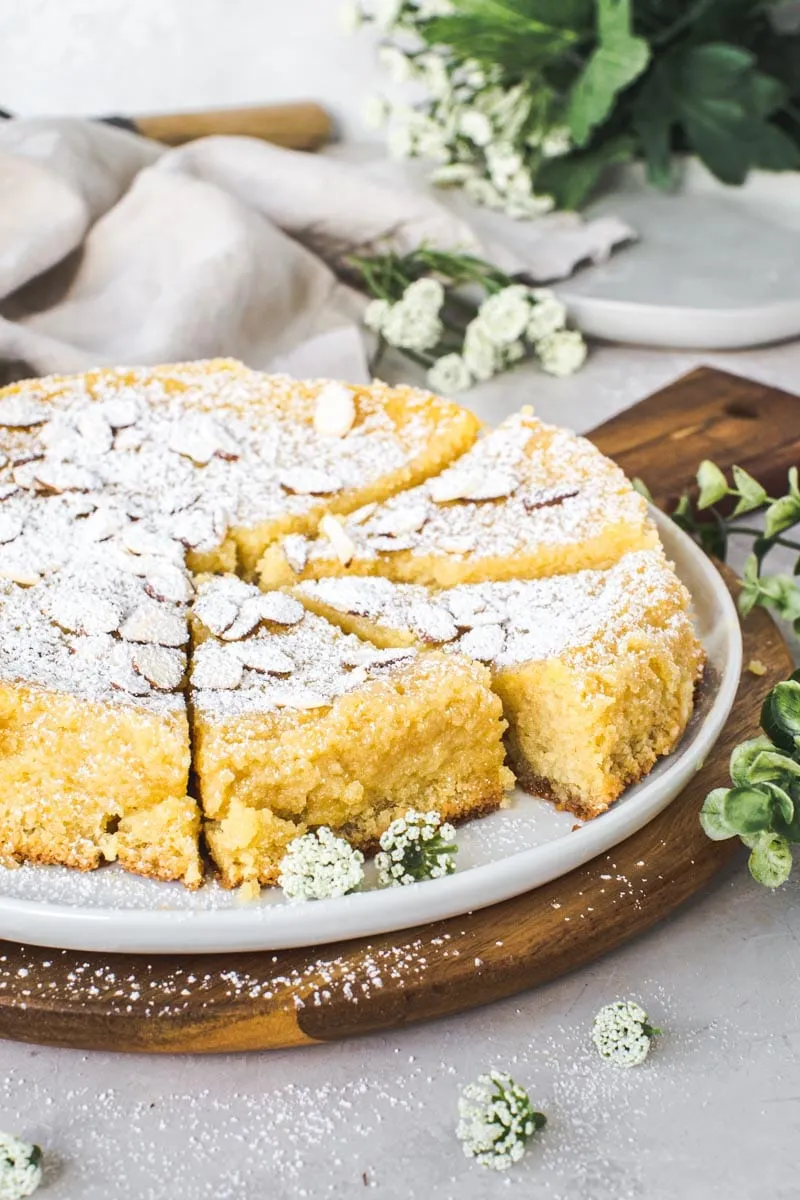 Lemon ricotta almond cake with slices cut out on a white plate with white flowers surrounding it.