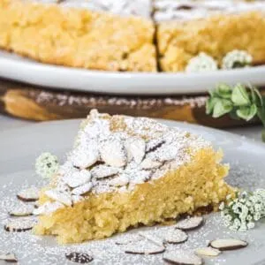 Slice of lemon ricotta almond cake on a white plate with almonds surrounding it and powdered sugar dusted on top.