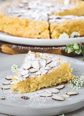 Slice of lemon ricotta almond cake on a white plate with almonds surrounding it and powdered sugar dusted on top.