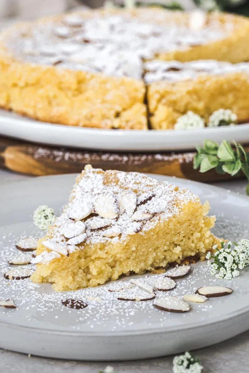 Slice of lemon ricotta almond cake on a white plate with almonds surrounding it and powdered sugar dusted on top.