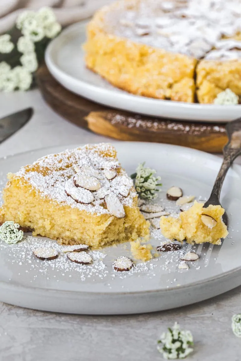 Slice of lemon ricotta almond cake with fork holding a bite sitting on a white plate.