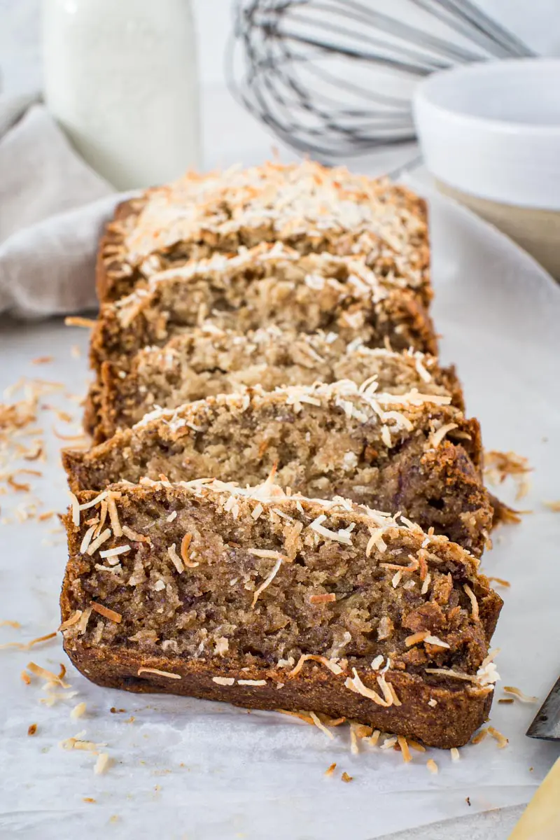 Slices of vegan banana bread topped with toasted coconut on wax paper.
