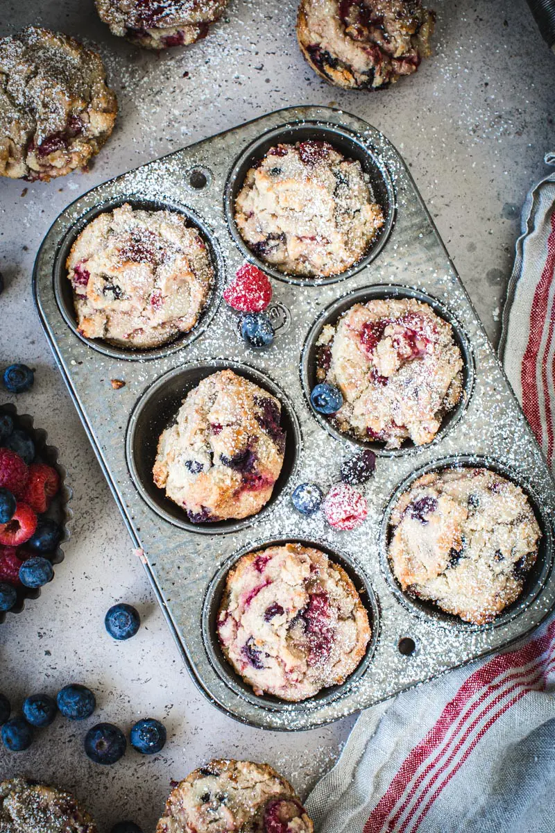 Powdered sugar coated mixed berry cornbread muffins in a muffin tin with muffins and berries surrounding it.