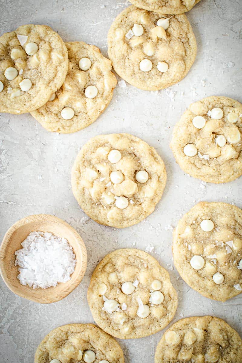 Salted white chocolate chip cookies spread out on a surface with a small bowl of salt flakes next to them.