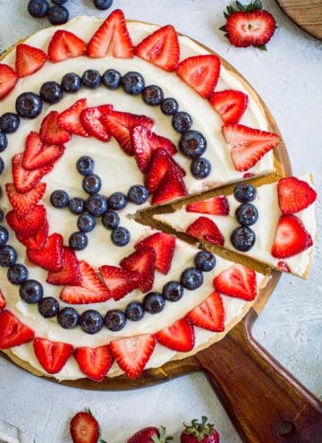 Fourth of July fruit pizza on a round wooden cutting board with strawberries surrounding it.